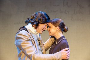 Two people on a theatre stage stand close together with their foreheads touching and arms around each other. One person wears a light-coloured historical-style coat and cravat, while the other wears a dark collared dress with hair tied back in a bun. Warm stage lighting illuminates the scene against a plain backdrop with faint handwritten text visible in the background.
