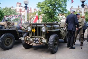 Colour photograph of two vintage military jeeps on display during an outdoor event, decorated with Union Jack bunting and flags. A group of people stand nearby in conversation, with trees and historic buildings visible in the background.
