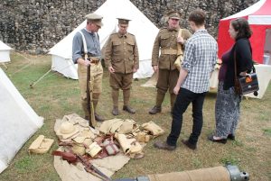 People speaking with First World War reenactors in period military uniforms at an outdoor encampment, with canvas tents behind them and historic equipment including rifles, packs and helmets laid out on the ground in front.