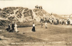 Sepia-toned photograph of a golf scene at Birkdale Links, Southport, with a player preparing to take a shot in the foreground and a large crowd gathered on sandy dunes in the background. Visible text reads: “Miss Dorothy Campbell on Birkdale Golf Links, Southport.”