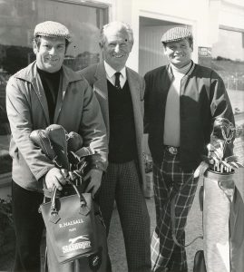 Black-and-white photograph of three people standing outdoors holding golf bags filled with clubs, dressed in vintage golfing attire including jackets, patterned trousers, and flat caps, with a clubhouse building in the background. A golf bag in the foreground reads “R. Halsall Plays Slazenger.”