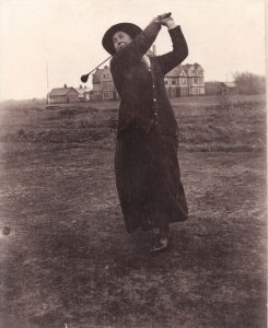 Sepia-toned photograph of a golfer in early 20th-century clothing mid-swing on a course, with large houses and open land visible in the background.