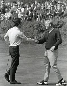 Black-and-white photograph of two golfers shaking hands on a putting green, with a crowd of spectators standing behind a fence watching the moment.