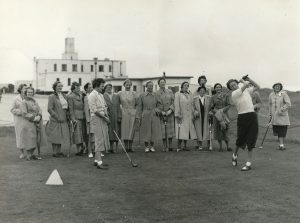Black-and-white photograph of a group of golfers standing on a course, watching as one player swings a club, with a clubhouse building visible in the background.