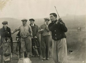 Black-and-white photograph of a golfer finishing a swing on a misty course, with several onlookers in caps and coats standing nearby beside golf bags.
