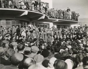 Black-and-white photograph of a large crowd gathered outside a modernist clubhouse, with people standing on balconies and around a podium where a large trophy is displayed during a public presentation.