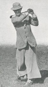 Black-and-white photograph of a golfer mid-swing on an open grassy course, wearing loose early 20th-century clothing and a hat.