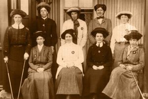 Sepia-toned group portrait of a women’s golf team seated and standing outside a building, all dressed in early 20th-century attire and hats, some holding golf clubs.