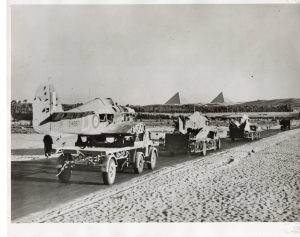 Black-and-white photograph of several vintage aircraft parked in a row on a grassy airfield, with their wings and propellers visible. Temporary structures and tents stand behind the planes, and open land stretches into the distance in the background.