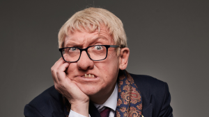 Studio portrait of a person wearing black-framed glasses, a dark suit jacket, white shirt, patterned scarf, and burgundy tie. The person rests one side of their face on a hand and looks directly at the camera. The image is tightly cropped against a plain grey background with even studio lighting.